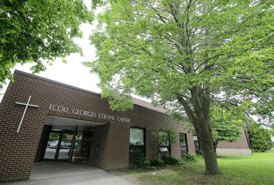 L'entrée de l'École élémentaire catholique George-Étienne-Cartier, une façade de brique brune surmontée d'une croix, à l'ombre d'un grand arbre.