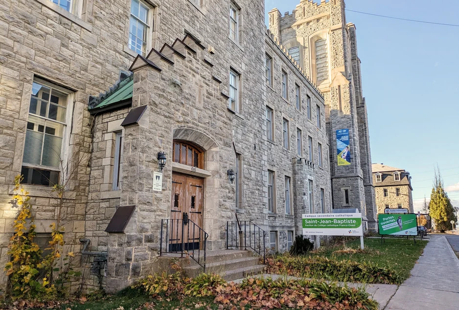 L'édifice de pierre qui abrite l'École secondaire catholique Saint-Jean-Baptiste, avec sa porte de bois en arche et sa tour crénelée à l'arrière-plan.